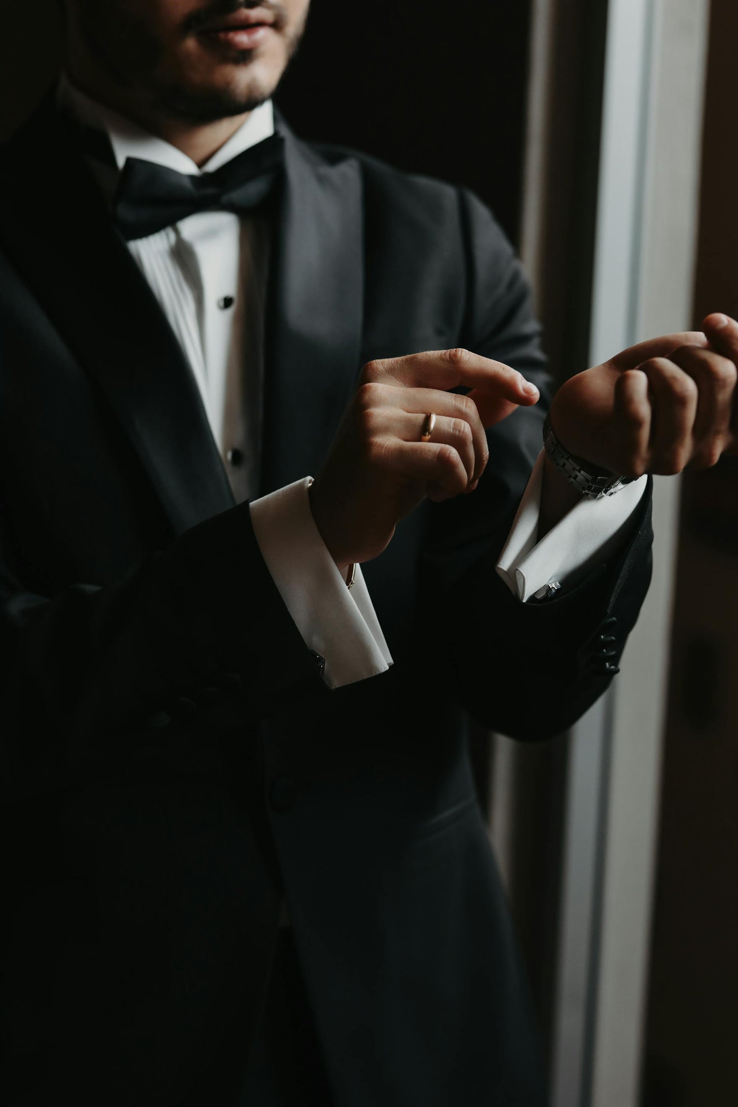 Man in a black tuxedo adjusting his jacket in a dimly lit room.